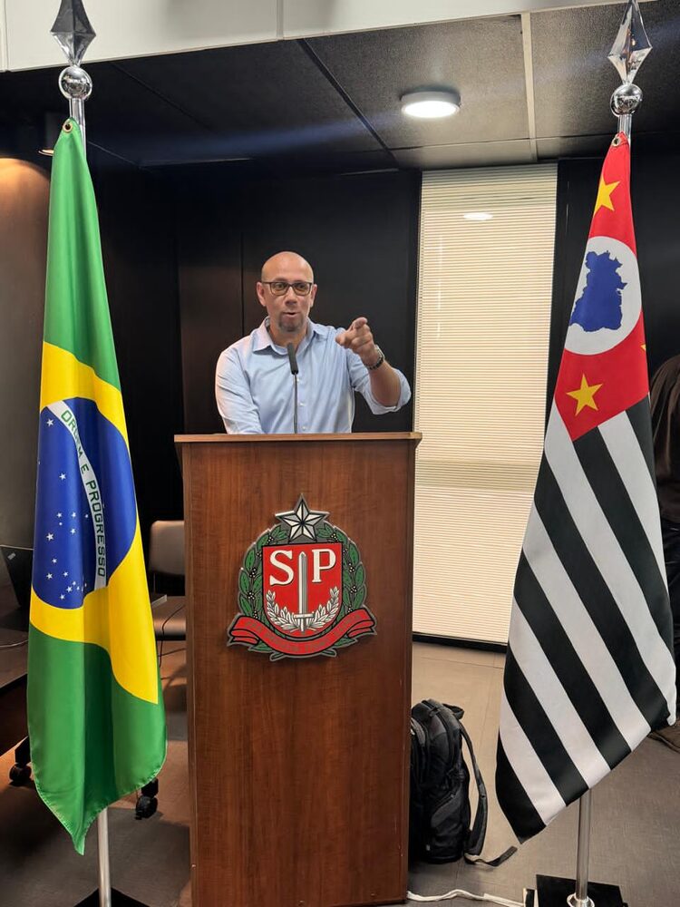 Rodrigo Moraes discursando em tribuna, entre a bandeira do Brasil e a de São Paulo